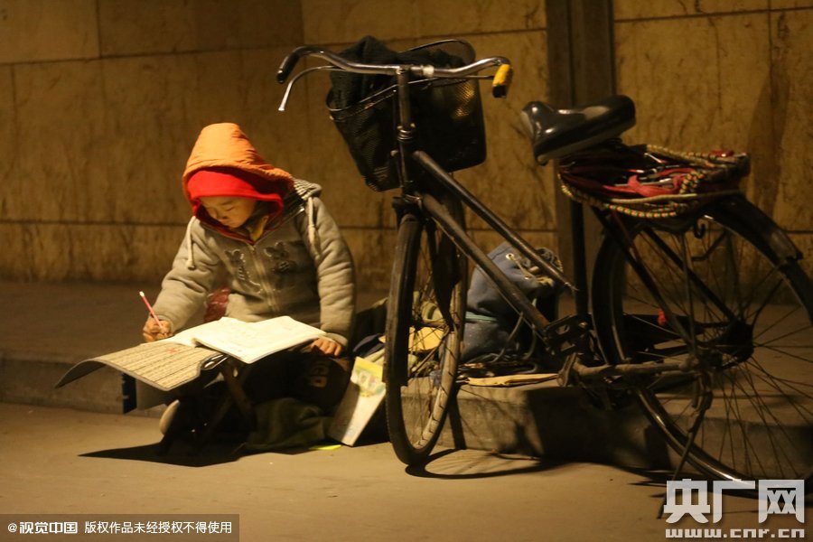 鄭州中原路京廣鐵路大橋下涵洞的馬路牙子邊，一位戴著紅色辣椒帽的小男孩趴在一個小馬扎上，上面鋪著一張車墊，在微弱的燈光下寫作業(yè)