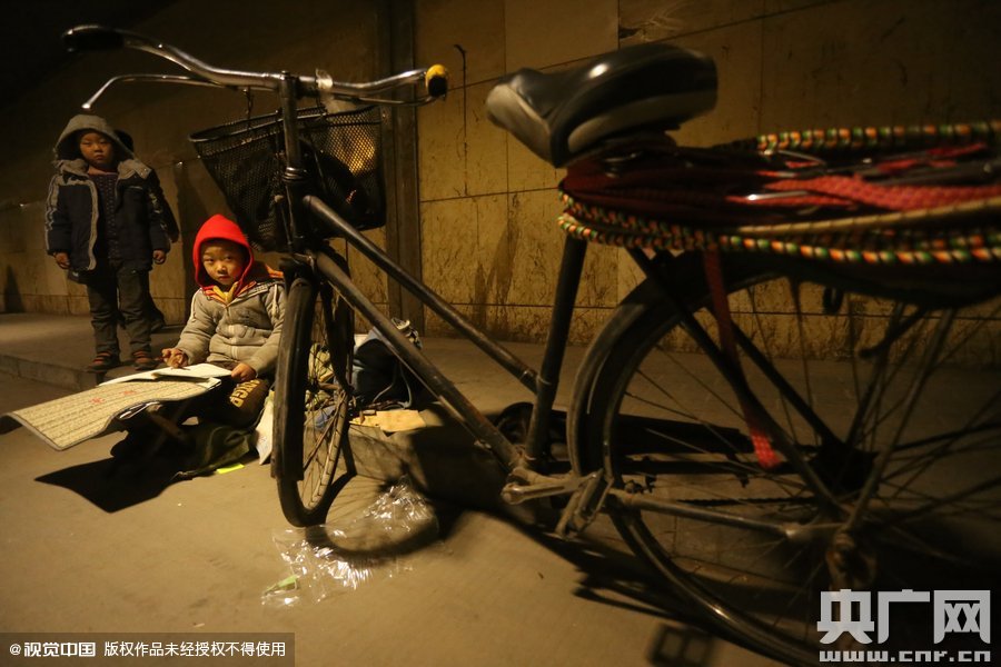 2015年11月8日晚，鄭州，7歲男孩涵洞橋下陪父母練攤，路邊燈下坐小馬扎上寫作業(yè)。